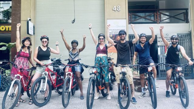 Seven people with bicycles pose together, smiling and raising one arm on a Hoi An city sidewalk in front of a building.