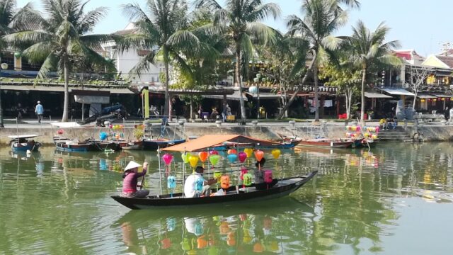 A small boat with colorful balloons floats on a river in Hoi An, with palm trees and buildings in the background.