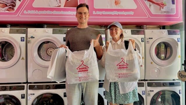 Two people smile in front of washing machines at a Hoi An laundromat, holding laundry bags beneath a pink Laundry Process sign.