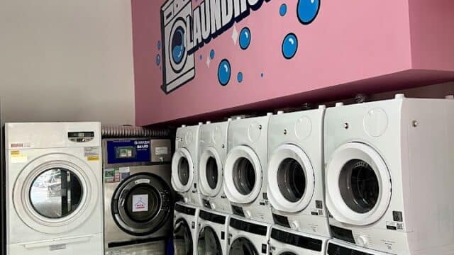 A laundromat in Hoi An with white washing machines and dryers against a pink wall, blue bubbles, and a classic Laundromat sign.
