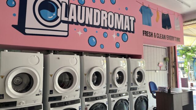 Row of washing machines in a Hoi An laundromat with a pink LAUNDROMAT sign and clothes hanging illustrations above.