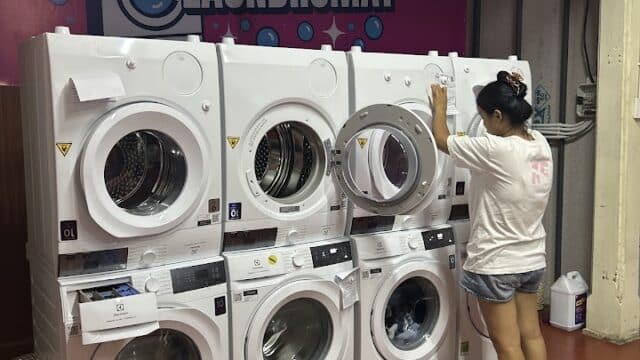 A person loads laundry into a washing machine at a Hoi An laundromat; a red basin with clothes sits on the floor.