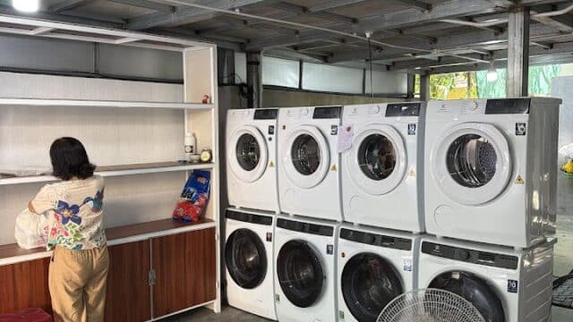 A woman stands by a counter near stacked washing machines and a fan in a Hoi An laundry room.