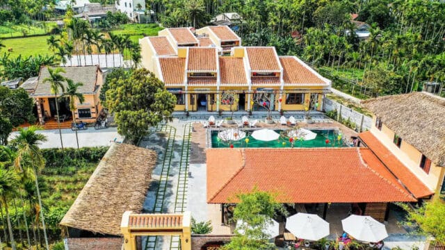 Aerial view of a Hoi An villa complex with a pool, lush greenery, red roofs, and outdoor seating areas shaded by umbrellas.
