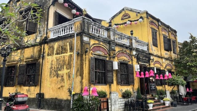 A yellow colonial-style building in Hoi An with wooden shutters, pink lanterns, and outdoor seating on a cloudy day.