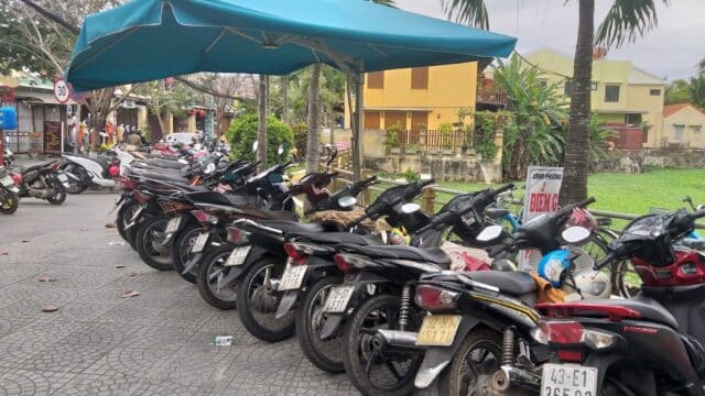 Rows of parked motorbikes under a blue canopy in Hoi An, with palm trees and yellow buildings in the background.