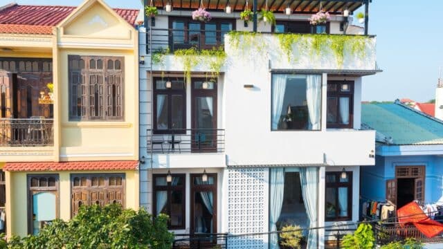 Modern three-story white house with large windows, balconies, and hanging plants, nestled between two traditional Hoi An houses.