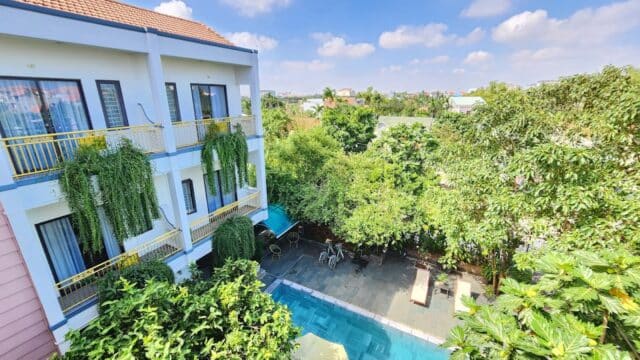 A bright, sunny view of a Hoi An hotel pool area with greenery, lounge chairs, and a two-story building with balconies.