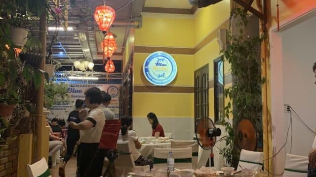 Indoor restaurant scene in Hoi An with round tables, white chairs, red lanterns, plants, and people dining under warm lighting.