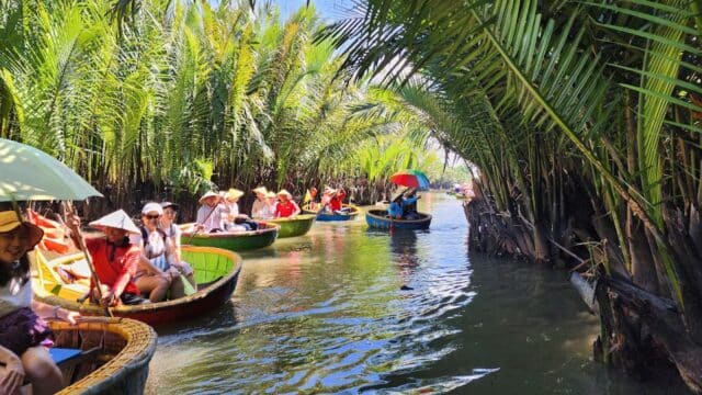 Tourists in round boats paddle through Hoi An’s narrow waterway, lined with lush palm trees under bright sunlight.