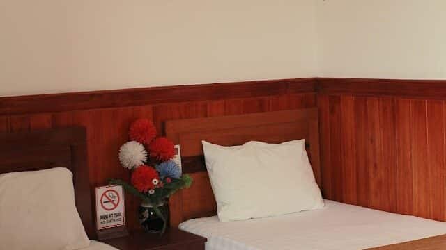 Two single beds with brown covers, white bedding, a nightstand, and a framed Hoi An cityscape on the wall.