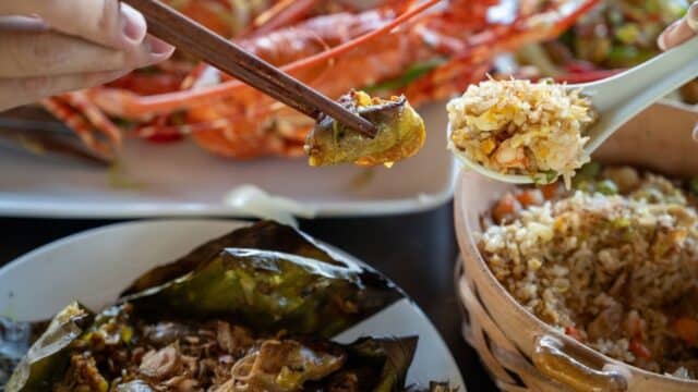 Chopsticks hold food near a plate of lobster in Hoi An, with fried rice above a bowl and wrapped dish in the foreground.