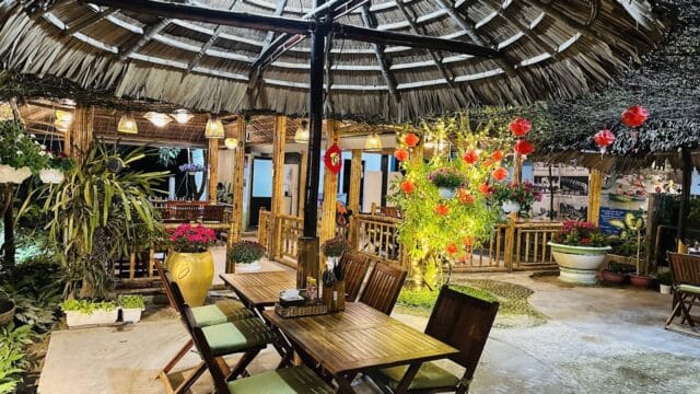 Outdoor dining area with wooden tables, plants, and lanterns under a thatched roof, evoking the charm of Hoi An at night.