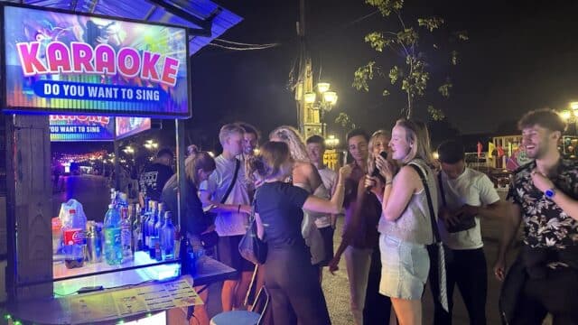 A group of people sing and laugh at an outdoor karaoke booth in Hoi An at night, under bright lights.