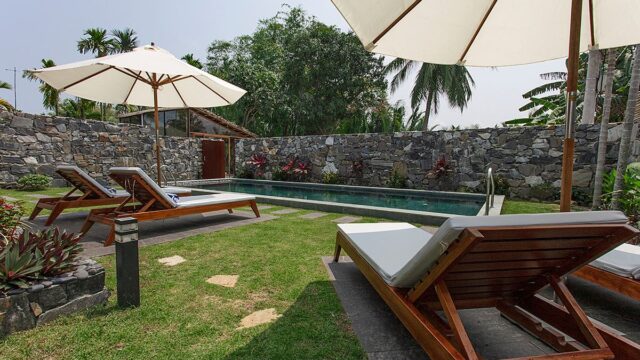 Lounge chairs and umbrellas surround a small outdoor pool in a Hoi An garden with stone walls and tropical plants.