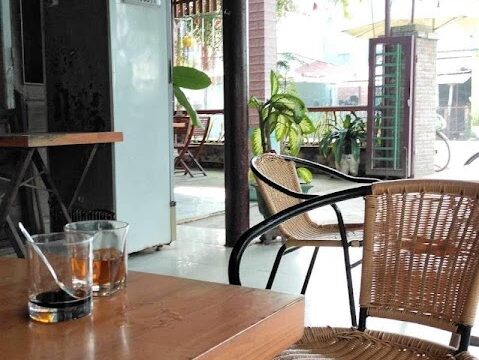 A wooden table with two glasses, wicker chairs, and a thatched roof in a Hoi An open-air cafe with plants and hanging red lanterns.