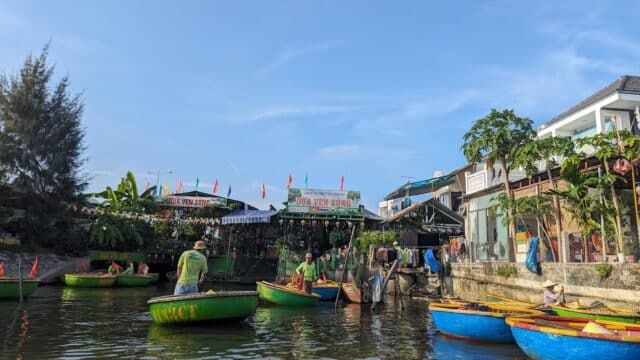 Round boats with people float on a river in Hoi An, near riverside buildings and greenery under a clear blue sky.
