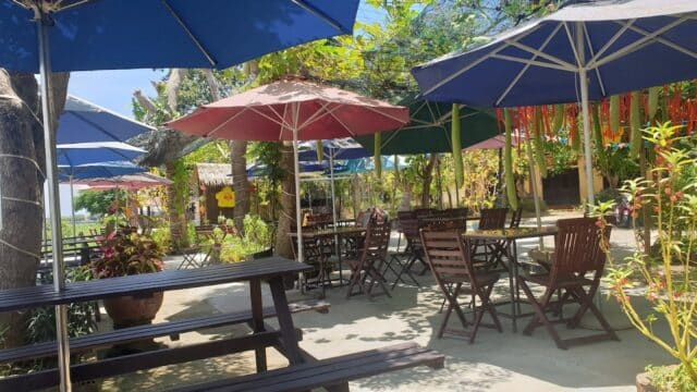 Outdoor café in Hoi An with empty wooden tables and chairs under large umbrellas, surrounded by greenery on a sunny day.