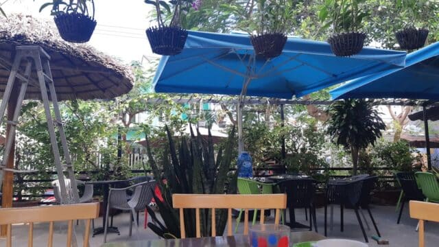 Outdoor café in Hoi An with empty chairs, hanging plants above, and a table featuring an empty cup and colorful glass in the foreground.