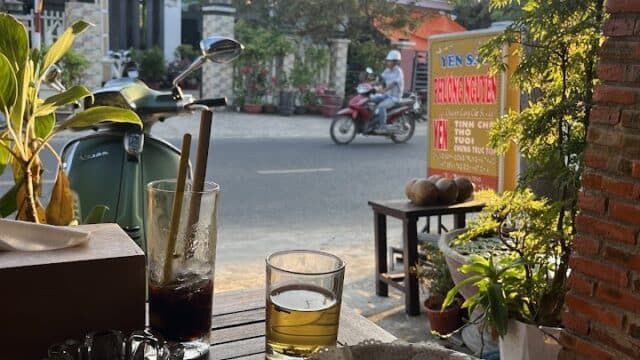 A meal with sandwiches, fries, and drinks on an outdoor table in Hoi An, with houses and a motorbike passing by across the street.