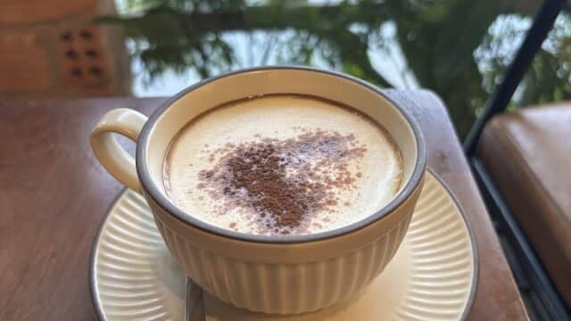 A cup of cappuccino with cocoa powder rests on a saucer by a window, overlooking Hoi An’s lush greenery.
