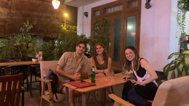 Three people sit at an outdoor cafe table in Hoi An at night, smiling, with drinks and lush greenery surrounding them.
