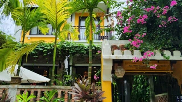 Yellow building with balconies in Hoi An, surrounded by palm trees, flowering plants, and a black entrance gate.