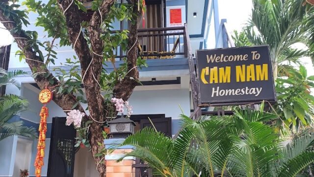 Entrance to Cam Nam Homestay in Hoi An with lush greenery, lanterns, and a welcoming sign by the gate.