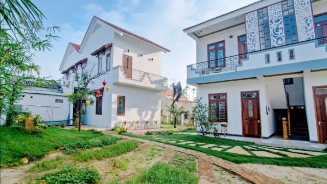 Two modern white houses with balconies in Hoi An, green lawns, and a stone path under a bright blue sky.