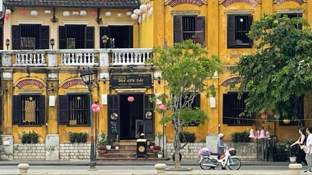 Yellow colonial-style buildings with lanterns by a river in Hoi An; people walk and a child rides a bike along the street.