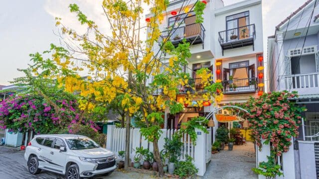 A white house with balconies in Hoi An, yellow flowers, red lanterns, and a white SUV parked by the sidewalk.