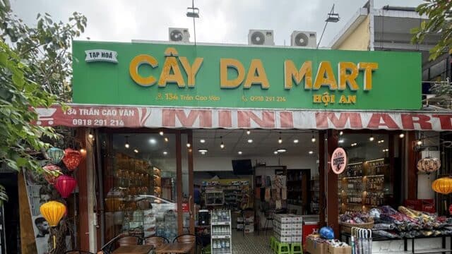A green sign reads Cây Da Mart Hội An above a mini mart storefront in Hoi An, with lanterns and goods displayed outside.