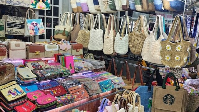 A market stall in Hoi An displays woven bags, wallets, and colorful souvenirs under hanging lights.