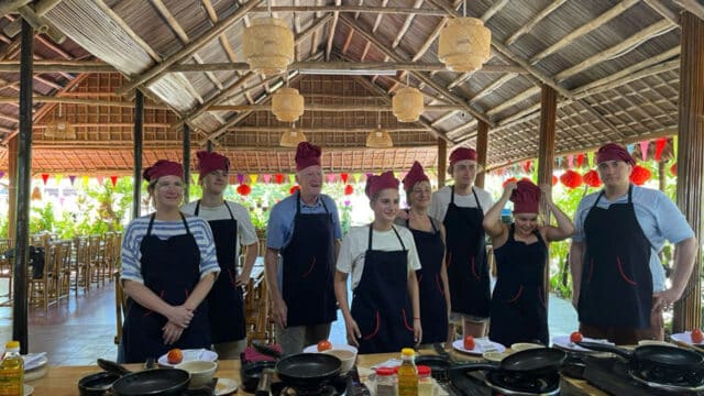 Eight people in aprons and chef hats stand in a rustic, open-air Hoi An kitchen with cooking stations and utensils.