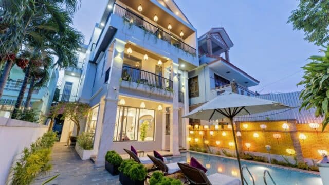 Modern three-story Hoi An house with poolside lounge chairs, large umbrella, and warm outdoor lighting at dusk.