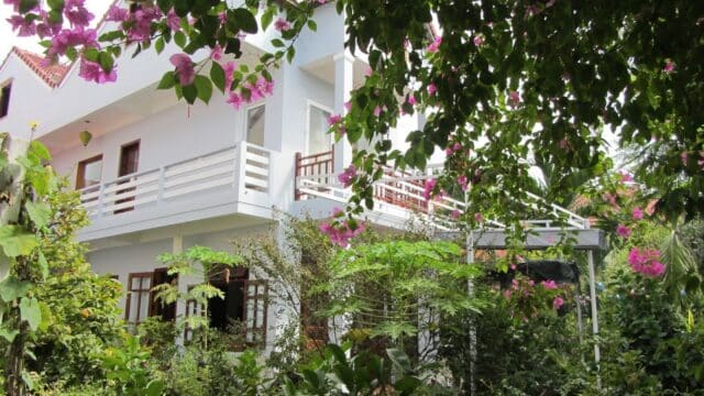 White two-story house in Hoi An, surrounded by lush green plants and pink flowers, viewed through leafy branches.