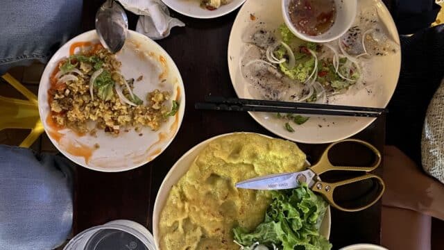 A table in Hoi An with partially eaten food, fresh vegetables, dipping sauce, chopsticks, and scissors on plates, viewed from above.