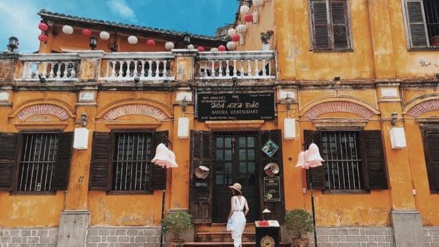 A woman in a white dress stands outside a yellow, lantern-adorned Hoi An building with wooden shutters and a sign above the door.