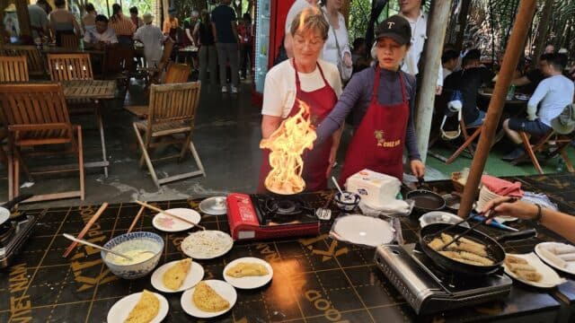 Two women cooking at a Hoi An food stall with a flame on the stove, surrounded by dishes and people dining in an outdoor setting.