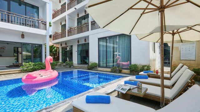 Modern Hoi An hotel pool with white loungers, blue towels, a pink flamingo float, and white umbrellas on a sunny day.