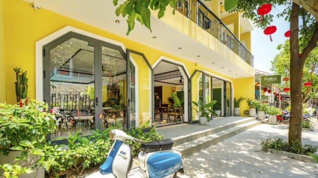 A yellow Hoi An building with large windows, lush green plants, and a blue-and-white scooter parked outside on a sunny day.