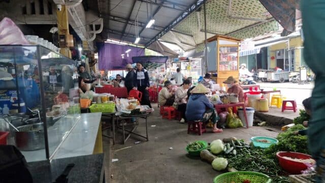 People shop and sell vegetables at Hoi An’s busy, covered market filled with food stalls, tables, and fresh produce on the ground.