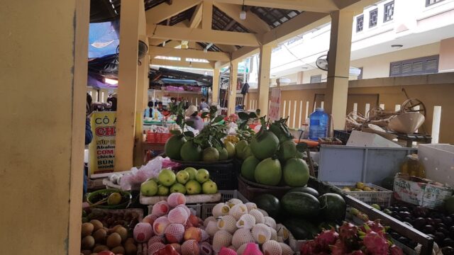 Hoi An market stall with apples, watermelons, pomelos, and dragon fruit displayed under a bright yellow roof.