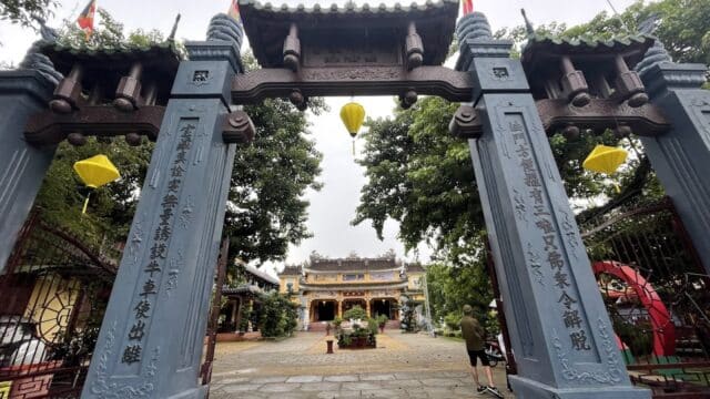 Large ornate entrance gate with pillars, lanterns, and a temple visible in the background, evoking the charm of Hoi An, trees on both sides.