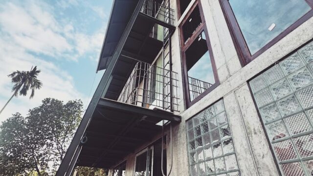 A modern Hoi An building with large glass windows, concrete walls, and plants outside, viewed from a low angle against a blue sky.
