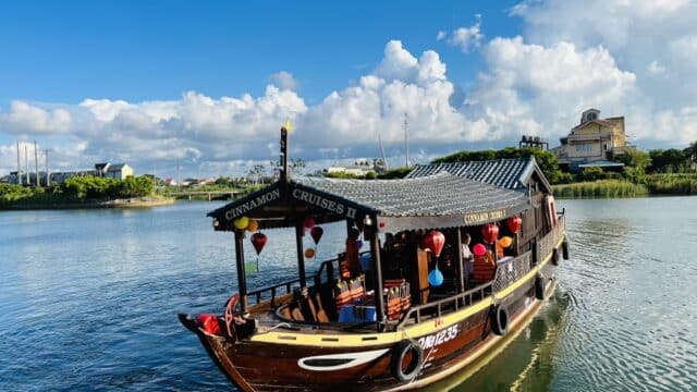 A wooden tour boat decorated with lanterns sails on Hoi An’s calm river under a bright blue sky with scattered clouds.