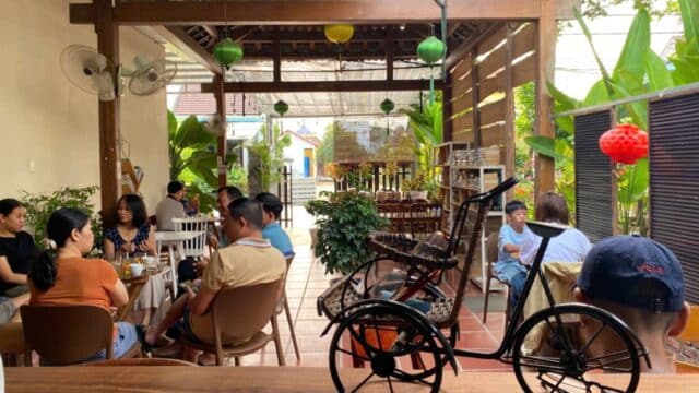 People are dining and chatting in an open-air Hoi An café with plants, wooden decor, and a tricycle model on a table in the foreground.