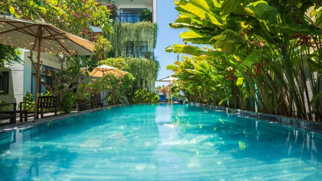 A clear blue swimming pool surrounded by lush tropical plants and sun umbrellas at a modern Hoi An hotel.