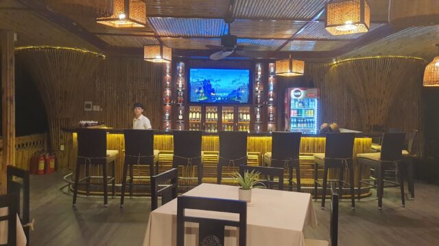 A bartender stands behind a bamboo bar with high stools in cozy, Hoi An-inspired, tropical-themed décor, warm lighting, and a TV.
