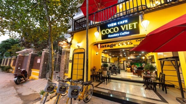 Bright yellow Hoi An café exterior with bikes parked outside, red umbrellas, and outdoor seating under warm lights at dusk.
